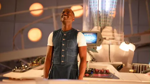 Nucti Gatwa is in front of a desk in the Tardis. He is wearing a striped denim top and skirt.