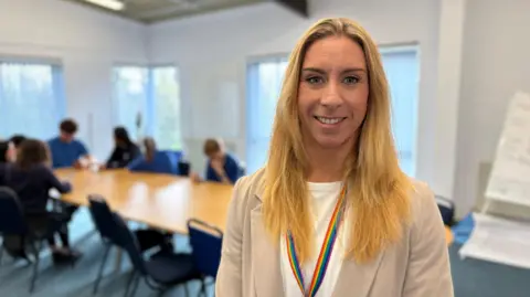 Katherine Ogden smiles. She is dressed in a light beige jacket and white T-shirt. There is a multicoloured lanyard around her neck. There are several pupils sitting at desks in the background.