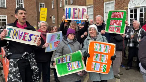 A group of people, some with learning disabilities, hold home made banners at the demonstration. The banners read 'invest in social care', 'help' and 'social care changes lives'. 