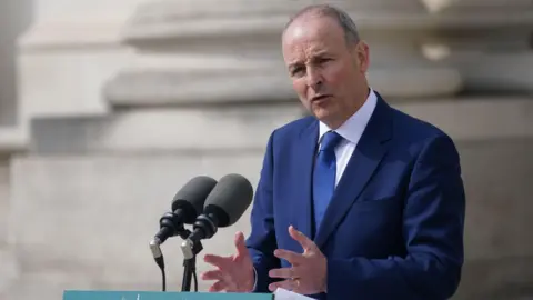 Micheál Martin is dressed in a blue suit, with a white shirt and navy tie. He is standing at a lectern. Two microphones are in front of him 