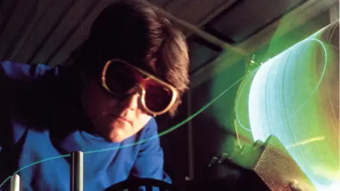 A photo taken in the 1980s of an engineer working on optical fibre. He wears large protective goggles and a blue jumper. He has brown hair. Fibre wrapped around a cylinder glows a green colour as it is used within a machine.