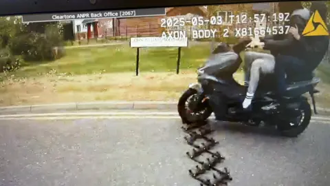 A still from a police body camera of the moped riders travelling over the stinger trap. The front wheel is directly above the spikes. There are two people on the moped, one in control and another holding on to him from behind.