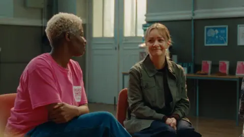 Breast Cancer Now Two women sitting in a room, smiling at each other. One of them is wearing a pink T-shirt with the logo of Breast Cancer Now.