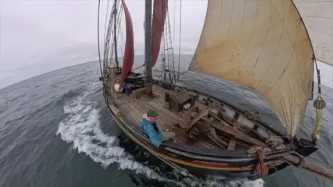 A large wooden ship is sailing on water. The boat is painted black on the outside, and has a beige coloured sail. 