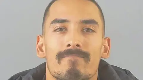 Hampshire Police A young man, with black cropped hair and a black moustache and beard stares directly at the camera. It is a mugshot taken by police. 
