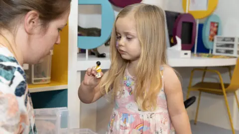 UCLH Seren is in the hospital play room, holding up a Lego-like toy figure. She wears a pink dress with clouds and rainbows on it. 