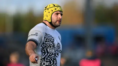 Bristol Bears Rugby player Will Capon during a match wearing a white shirt with black sponsor logos and a protective yellow cap on his head.
