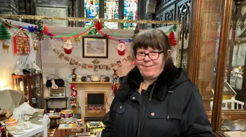 Rhonda Styles in black coat and wearing glasses stands in front of a exhibit at st columbs cathedral. She has brown hair.