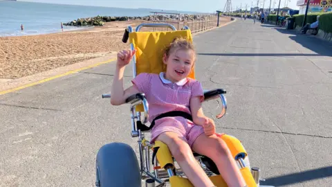 Jaimielee Rendall A young girl wearing a red-and-white school dress in a yellow beach wheelchair on the promenade.
