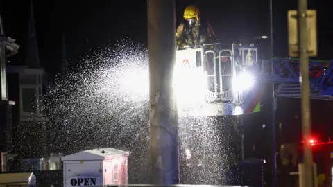 Pacemaker Press A fire fighter, wearing a hard hat and a mask, is in a lift off the ground putting out a fire on a mast. Sparks are flying around the mast. It's night.