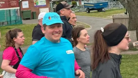 Ryan is running in a turquoise shirt and black shorts in the centre, surrounded by other participants wearing athletic clothing, with trees, fences, and buildings in the background.