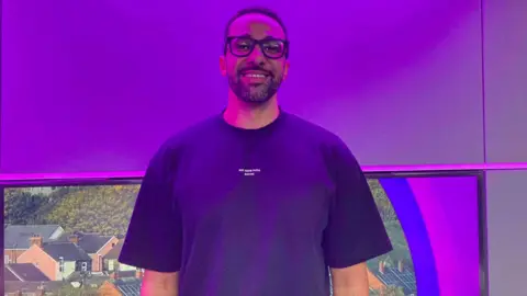 A man with glasses and a blue shirt stands in a studio and smiles while bathed in purple light.