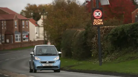 A blue car shown on a road with a road sign n the side displaying a 30 speed limit with the words 'Slow Down'.