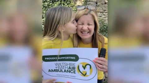 Klaire Alexander A woman holding a microphone and with a young girl giving her a kiss on the cheek while holding a sign which says 'Wiltshire and Bath Air Ambulance'