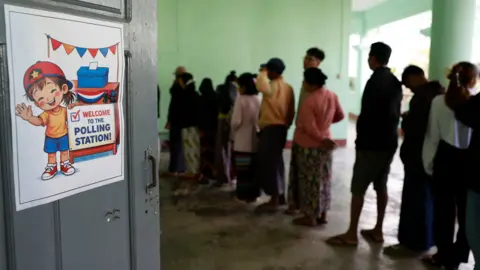 Myanmar voters queue up to cast ballots during the first phase of general election at a polling station in Naypyitaw, the capital city of Myanmar, 28 December 2025. The walls are green, and there is a poster of a smiling cartoon child saying "welcome to the polling station!"