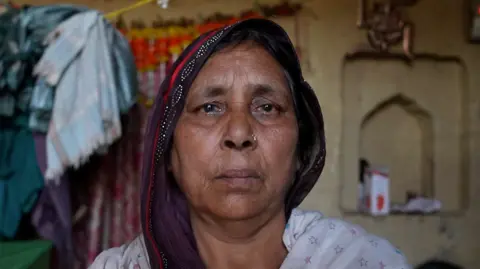 A woman wearing a purple saree with her head covered looks into the camera 