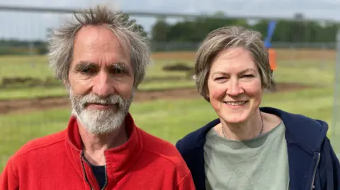 Luke Deal/BBC Angus Wainwright and Helen Geake stand side-by-side smiling at the camera in Sutton Hoo. Mr Wainwright has grey hair, a grey beard and is wearing a red fleece. Ms Geake has short grey hair and is wearing an olive green T-shirt with a navy hoodie over the top.