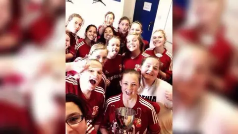 Swindon Town Community Foundation A joyous photo of a young women's football team in full kit in a changing room holding a trophy