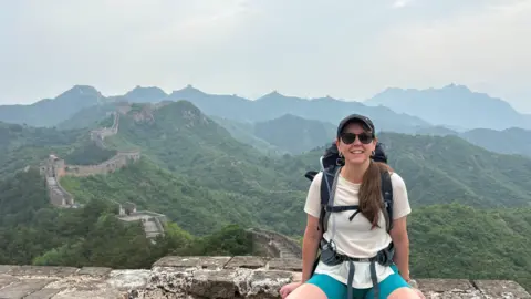 Ruth wears a white t-shirt and shorts and has a back pack on her ack.  She is sitting on part of the wall, which snakes through the hills in the background.