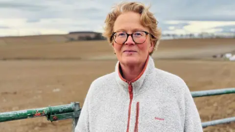 BBC Marguerite Fleming standing in front of a farm gate, she is wearing glasses and is looking at the camera, she is wearing a light grey fleece, and there are brown fields in the background.
