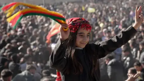 BBC, a child in the top black part and traditional blurring on top, waves a green, red and zero scarf while sitting on the shoulders of someone in the anniversary celebrations. A large crowd can be seen behind. 