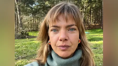 Katy Coe Katy Coe takes a selfie style image as she stands in a grass field with large trees behind her. She has blonde hair with a fringe. She is wearing a green turtle neck jumper and red and gold hoop earrings. 