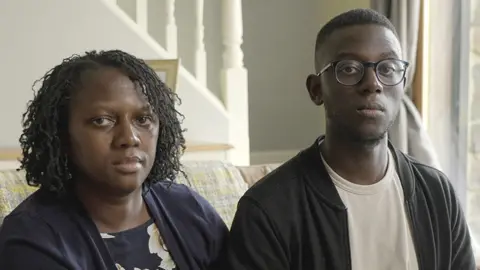 Celeste, a woman wearing navy, and Elias, a young man with glasses and looking at the camera and wearing a white T-shirt, sitting together on a sofa