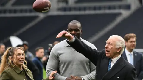 King Charles throws an American Football wearing a long coat and suit