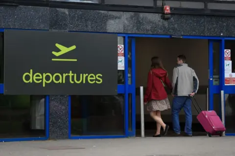 PA Media Woman wearing red coat and dress with man in grey jacket pulling red suitcase through blue sliding door with sign reading 'departures' beside it. 