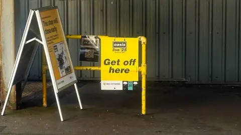 Emma Gibbs A sign reading Oasis Live '25 Get Off Here hangs off a yellow stanchion at a corrugated metal-built tram stop in Prestwich