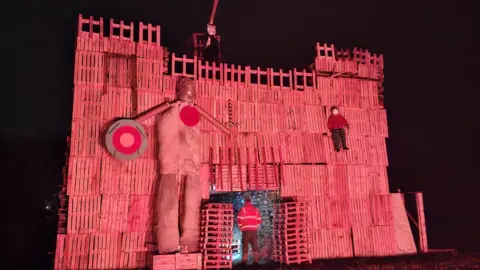 Colerne Bonfire Crew A fort shaped structure at least two storeys high made out of old pallets, with turrets and scarecrows. It is night time and the structure is light with a pinkish-red light.