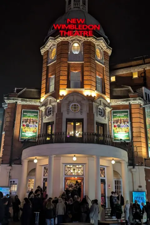A grand looking building lit up at night with crowds waiting outside. New Wimbledon Theatre signage sits on top of the building.