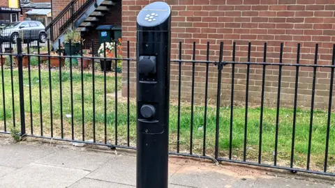 An electric car charging point next to a pavement, with a black fence, grass and a brick wall behind it. A car and shops can be seen in the background.