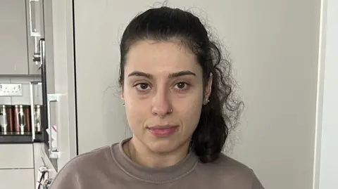 Emily Martins in her flat. She wears a taupe jumper.