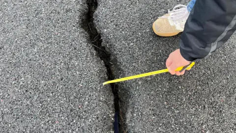 A close-up of a crack in a Tarmacked road, and a person is extending a yellow measuring tape into the crack.