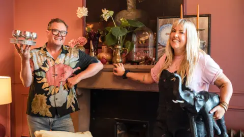 Alan Carr holding silver wine glasses and a woman holding an elephant statue in front of a fire place