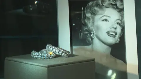 Sparkling earrings sit in a glass case in front of a portrait of Marilyn Monroe
