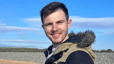 A man smiling at the camera. He is on a beach with sand and rocks behind him. He is wearing a coat with a fluffy hood. He has brown hair with a beard.