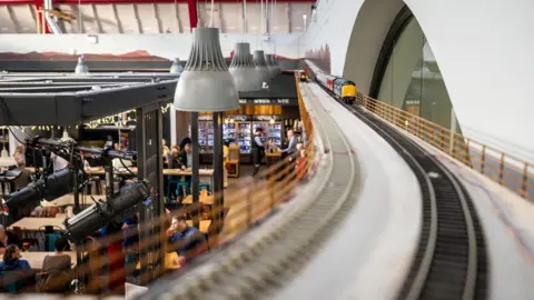 Paul Campbell Looking down a track towards the front of a yellow and blue locomotive, which is pulling carriages. Below the track are stalls in the Victorian Market.