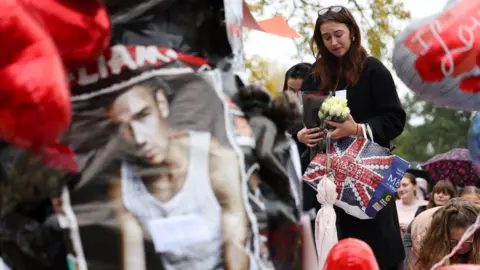 Reuters A woman looks down, eyes closed, as she holds two bouquets of flowers beside a memorial for Liam Payne with a picture of the singer and balloons