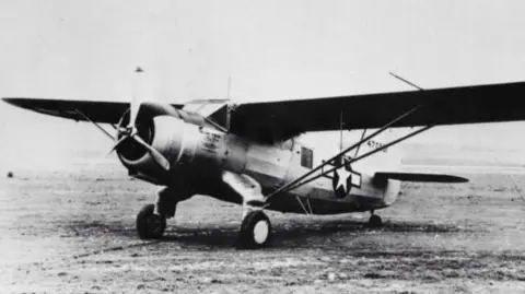 PA Media A black and white photo of the C-64 Norseman plane, sitting on a large field with nothing else around. It has one large flat wing spanning both sides over the top, a propeller at the front and two larger wheels below the front of the fuselage, and one small wheel at the back. 