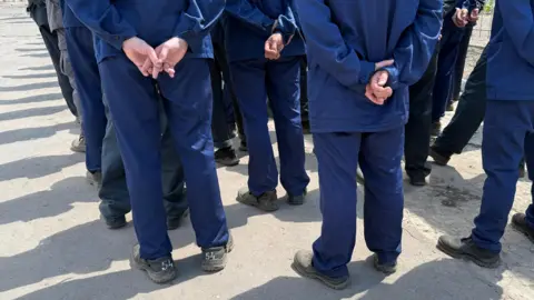 The backs of a line of men wearing a navy uniform. They are standing with one hand holding the other behind their backs. 
