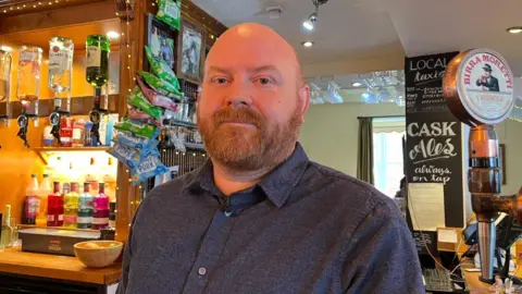 A bald man with a ginger beard stands behind the bar of a pub, optics and beer pumps can be seen in the background and foreground. He wears a navy shirt.