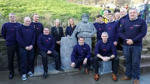 RNLI/Ceri Oakes Members of the RNLI Whitby crew around the wire sculpture, wearing blue jumpers and black trousers and all smiling