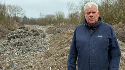 Graham Morgan has short white hair and a dark blue padded anorak. He stadns in front of a mountain of illegally tipped waste