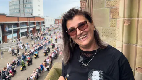 BBC Lili Myers wears a black t-shirt and sunglasses and smiles at the camera. She is standing on a balcony above Barrow's high street, where there is a procession of motorcyclists.