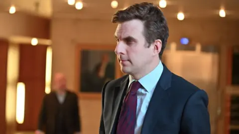 Getty Images Daniel Johnson, who has short dark hair, side on to the camera while in a brightly lit room. He is wearing a dark suit, light-blue shirt 