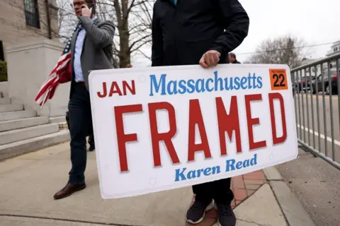 Getty Images A person holds a sign shaped like a Massachusetts license plate that reads FRAMED