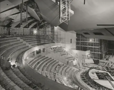 Sir Robert McAlpine/J E Rackham Photography Ltd Scaffolding fills a large unfinished interior space inside the National Theatre during construction. There is curved seating in tiers above a stage to the right that is being constructed. 