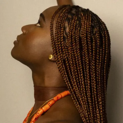 Rayh Mohammed A photograph of a woman with brown hair in braids. She is wearing an orange necklace and gold earring.